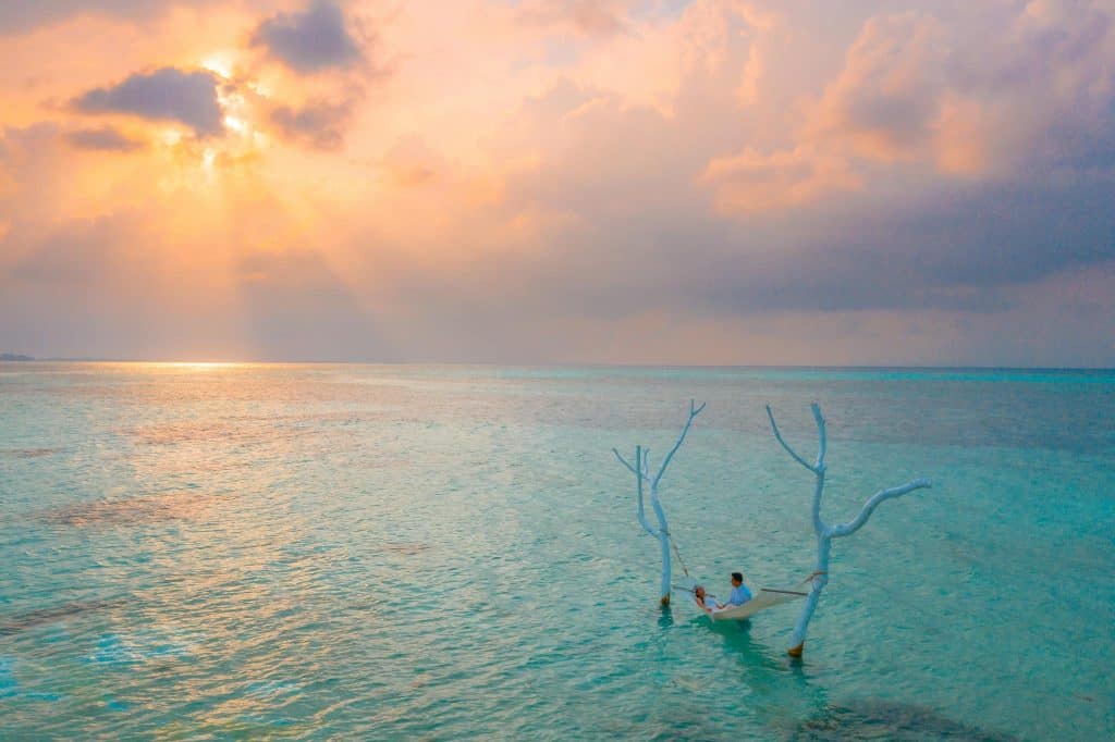 A couple relaxing in a hammock over the clear waters of Maldives at a gorgeous sunset.