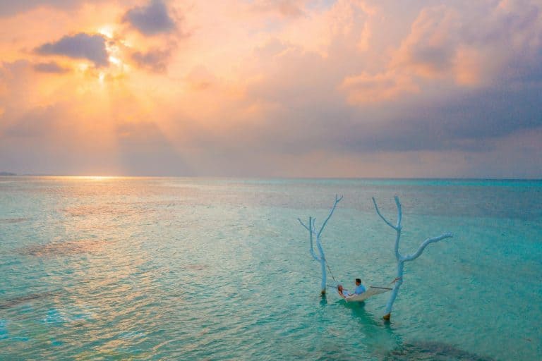 A couple relaxing in a hammock over the clear waters of Maldives at a gorgeous sunset.